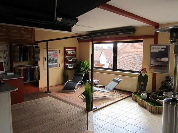 Moderner Ausstellungsraum mit Liegestühlen, Holzboden, Pflanzen und großem Fenster.